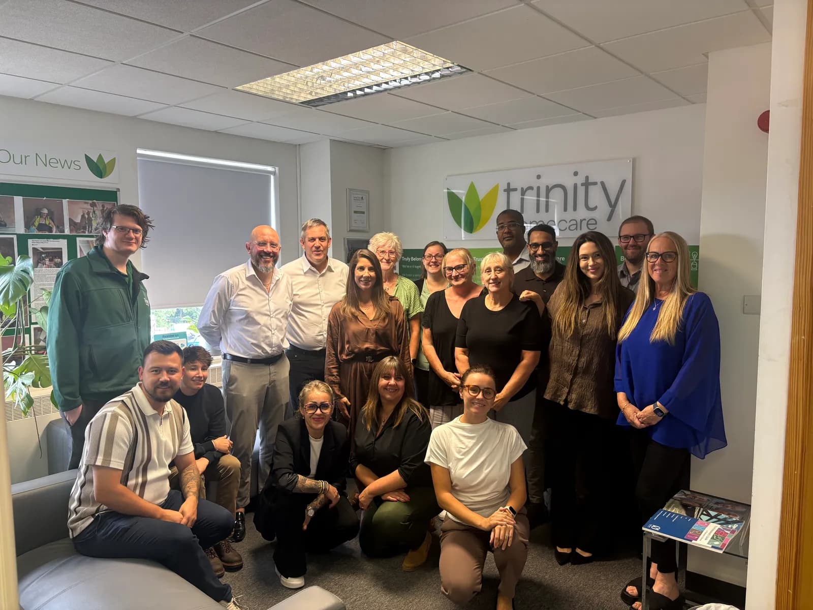 A diverse group of men and women of various ages are gathered for a group photo in an office setting, with some standing and others kneeling or sitting, beneath a "trinity healthcare" logo.